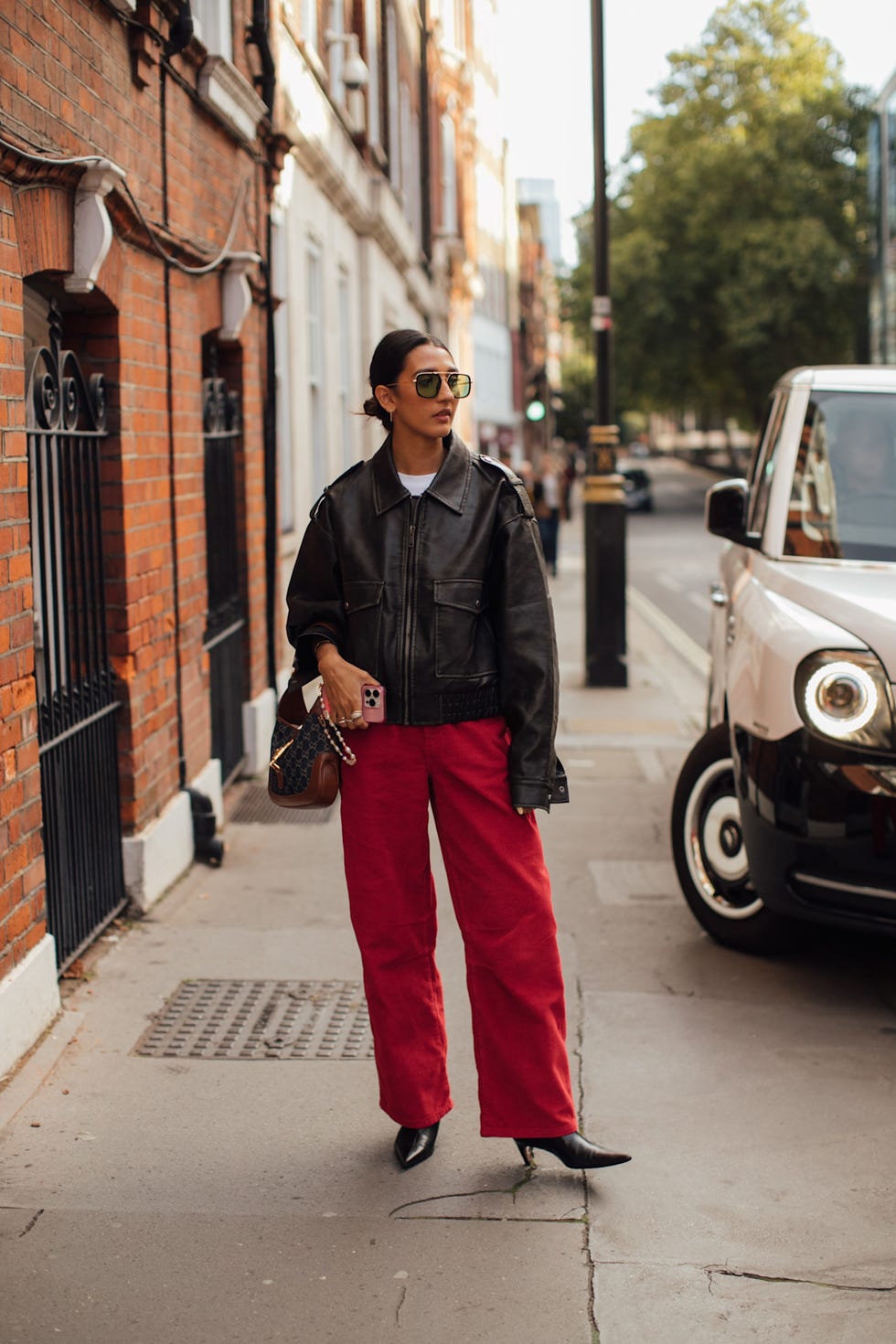 a person wearing a black leather jacket and red pants stands on a city street with a vehicle nearby